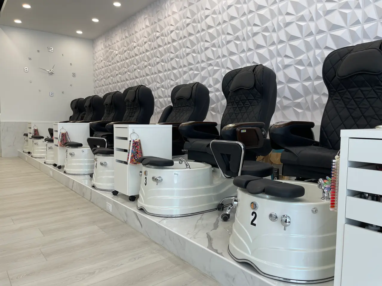 Row of black quilted pedicure massage thrones with white soak basins against a sculpted geometric feature wall at Aivia Nails in Cranbrook